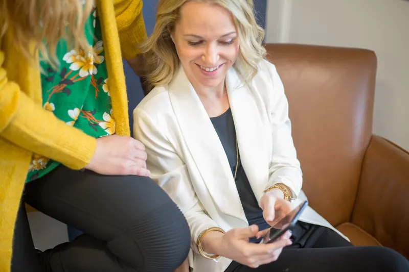 Dos mujeres sonrientes revisando juntas la pantalla de un teléfono móvil, una sentada en un sillón de cuero y otra de pie
