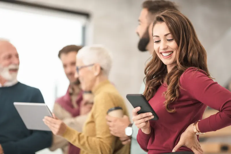 Grupo de personas conversando mientras una mujer sonriente usa su teléfono móvil y otros sostienen una tableta