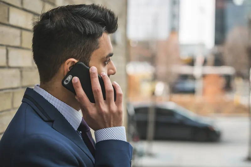 Hombre de traje azul hablando por teléfono móvil junto a una pared de ladrillo