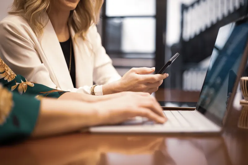 Manos de una mujer sosteniendo un teléfono móvil mientras otra persona escribe en una laptop sobre una mesa de madera