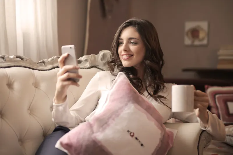 Mujer sonriente sentada en un sillón con un teléfono móvil en una mano y una taza en la otra