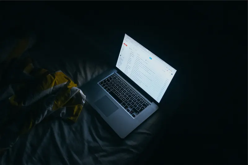 Laptop abierta sobre una cama en la oscuridad mostrando en pantalla una lista de mensajes de correo