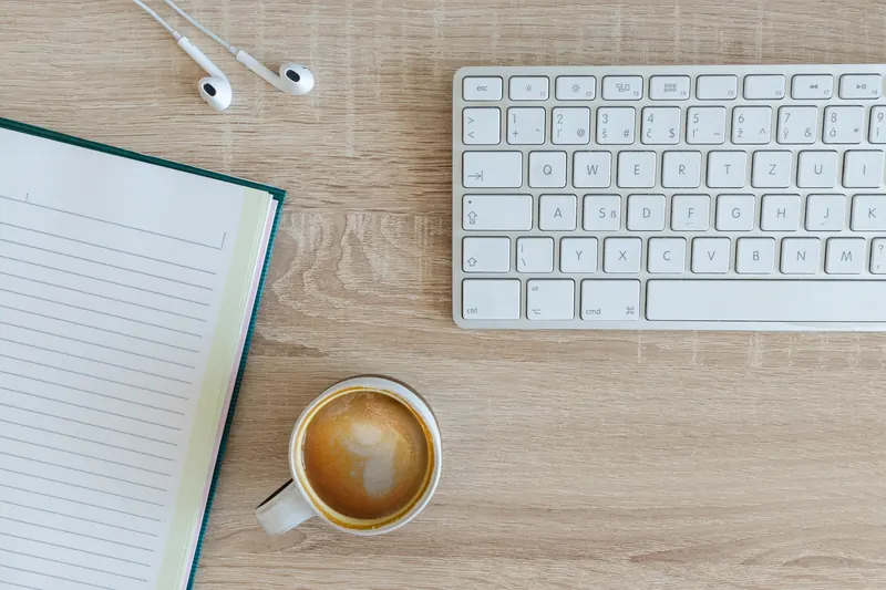 Teclado inalámbrico, cuaderno, audífonos y una taza de café sobre un escritorio de madera clara, vista desde arriba