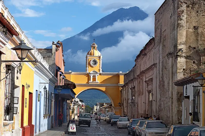 Calle de Antigua Guatemala con un arco amarillo con reloj sobre la calzada y un volcán al fondo