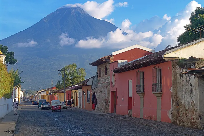 Calle empedrada de Antigua Guatemala con casas coloniales de colores y un volcán al fondo bajo nubes