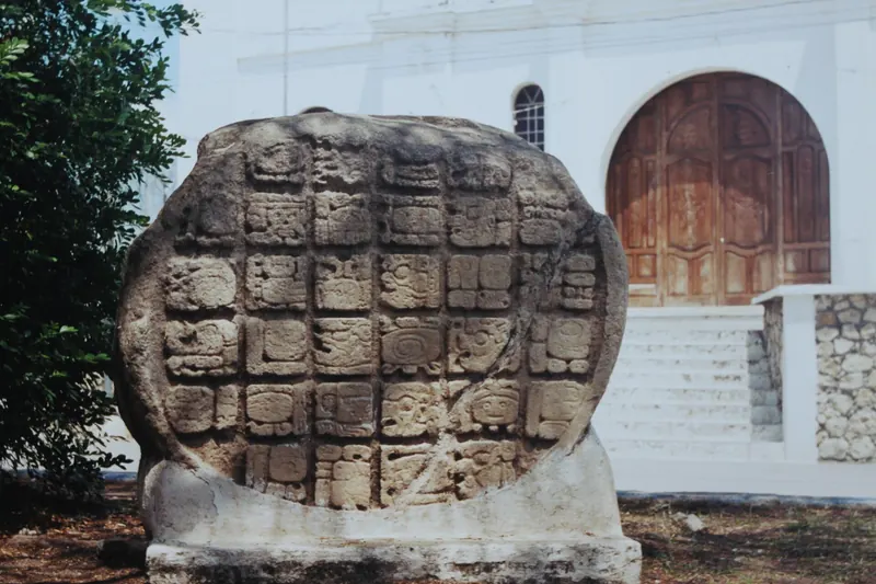Piedra maya redonda con glifos tallados frente a una iglesia blanca en Flores, Guatemala