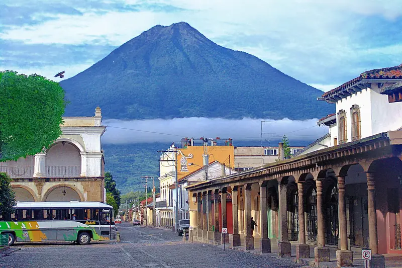 Portales coloniales de Antigua Guatemala con un autobús estacionado y un volcán asomando sobre las nubes