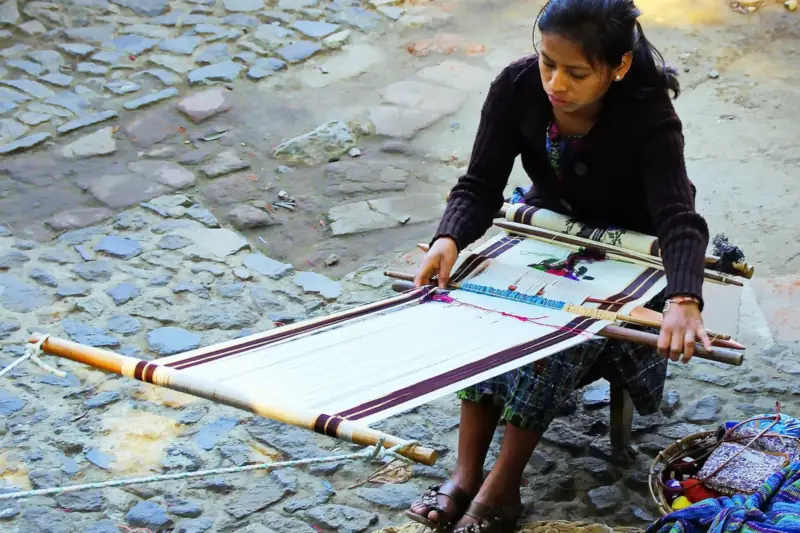 Mujer de Guatemala tejiendo en un telar de cintura sobre un piso empedrado, con telas de colores a un lado