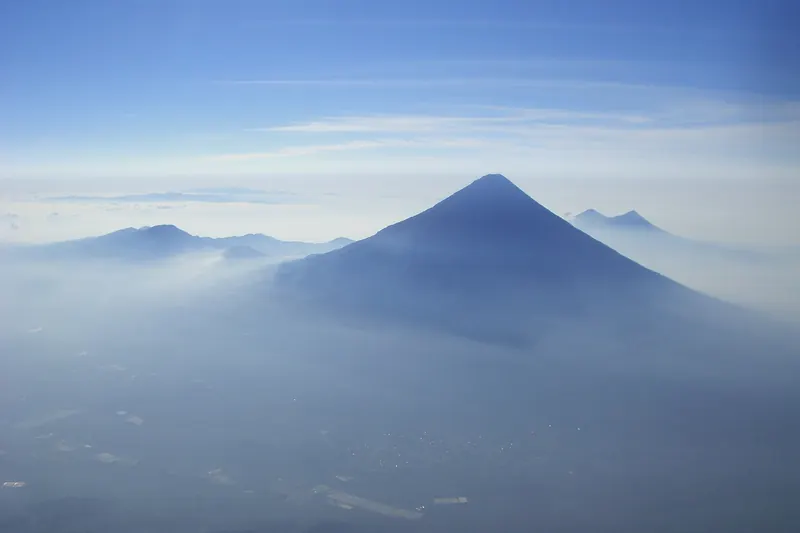 Volcán de forma cónica que sobresale de un mar de neblina bajo un cielo azul, visto desde las alturas