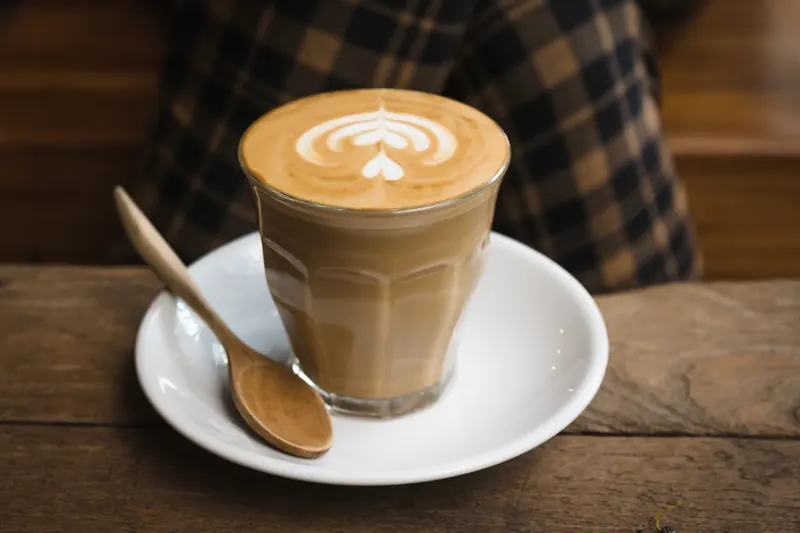 Café latte con espuma en un vaso de vidrio sobre un plato blanco con cuchara de madera