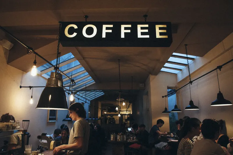 Interior de una cafetería con un rótulo luminoso de COFFEE, lámparas colgantes y clientes al fondo