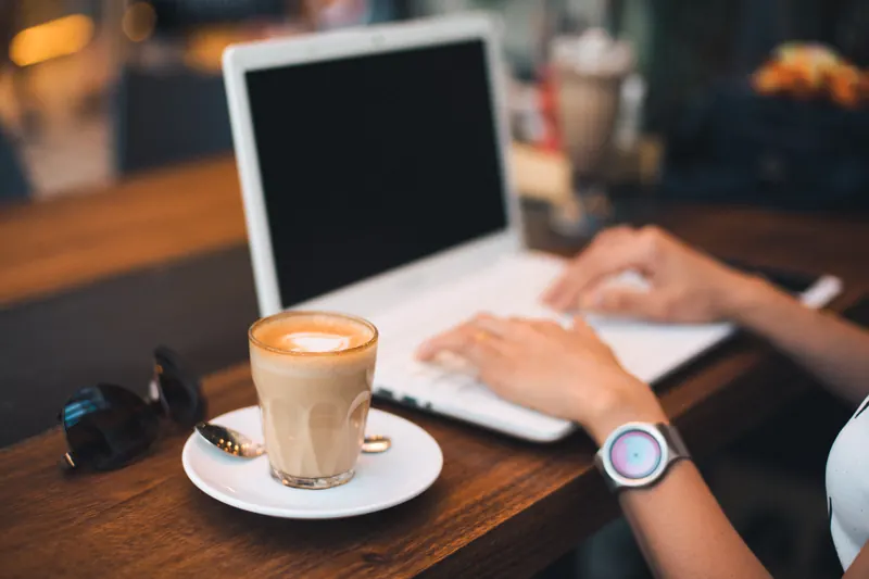 Persona escribiendo en una laptop blanca junto a un café con leche y lentes de sol sobre una mesa de madera