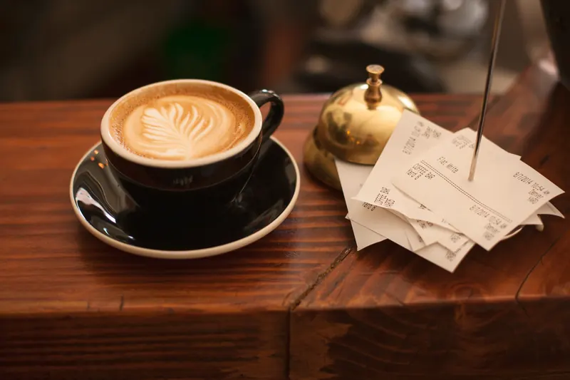 Taza de café con arte latte sobre una mesa de madera junto a una campana y recibos