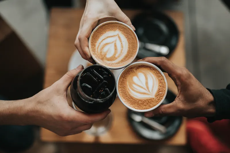 Tres manos acercando tazas de café latte sobre una mesa de madera, vista desde arriba