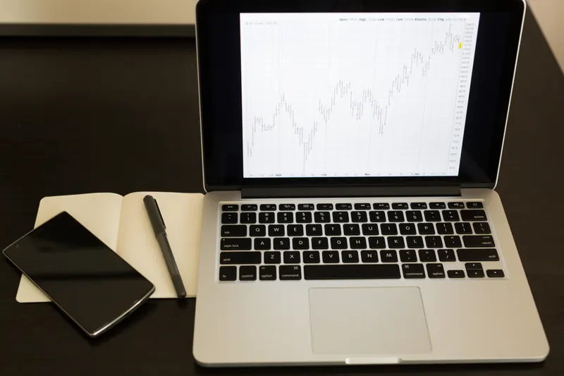 Laptop con una gráfica de líneas en pantalla, un cuaderno y un teléfono sobre una mesa oscura