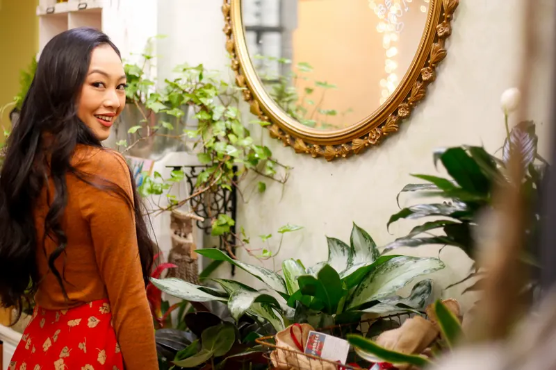 Mujer sonriente mirando hacia atrás dentro de una tienda con plantas y un espejo dorado en la pared