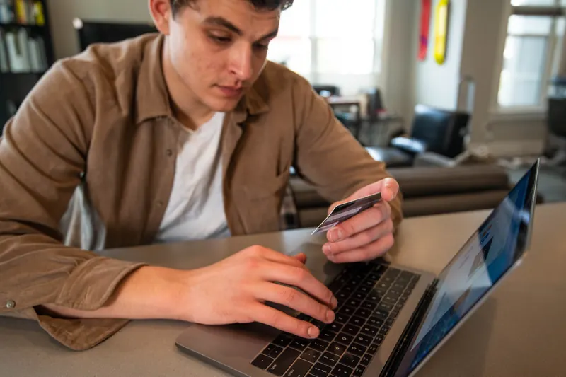 Joven de camisa café sosteniendo una tarjeta de crédito frente a una laptop para pagar en línea
