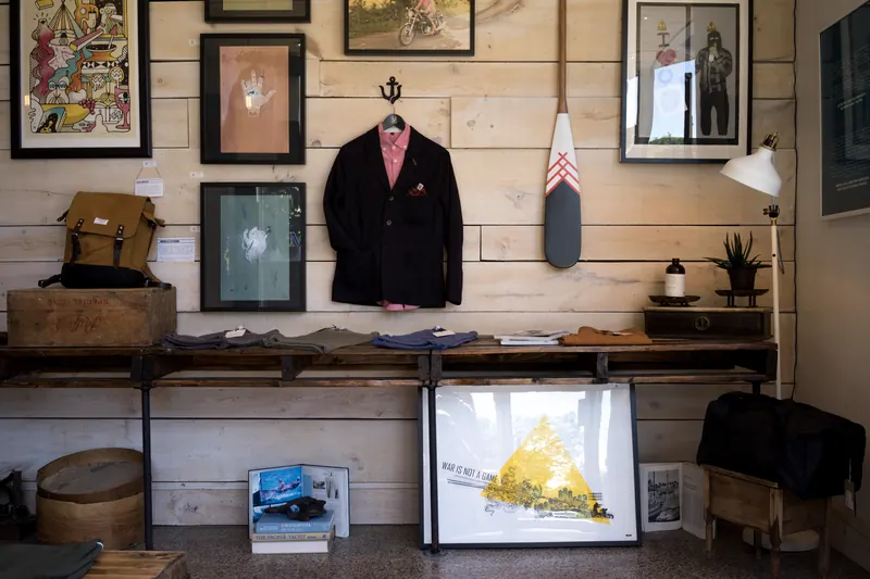 Pared de una tienda con un saco colgado, cuadros enmarcados y una repisa de madera con una lámpara
