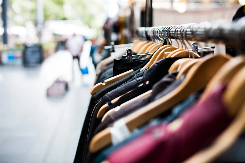 Ropa colgada en perchas de madera en un puesto de una calle comercial, con transeúntes desenfocados al fondo