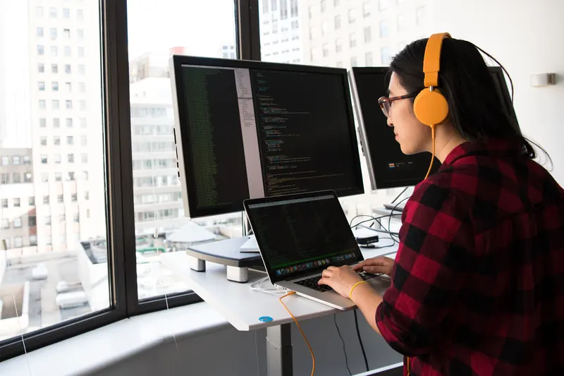 Mujer con audífonos naranjas programando frente a varios monitores junto a una ventana de oficina