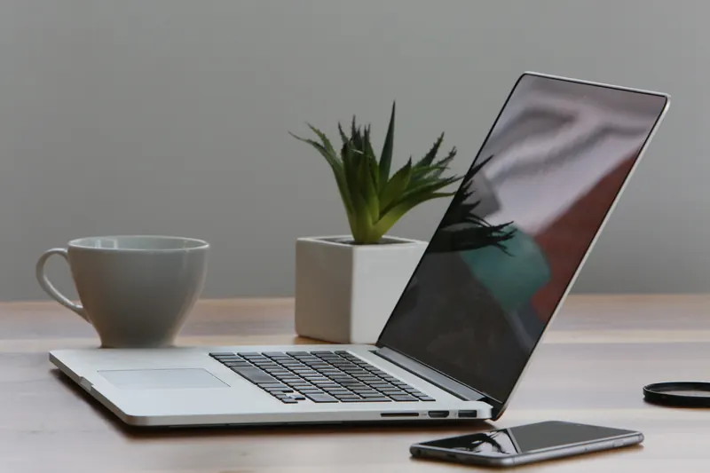Laptop abierta con una taza, una planta pequeña y un teléfono móvil sobre un escritorio gris