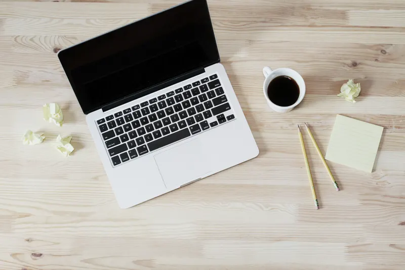 Laptop abierta sobre un escritorio de madera clara con una taza de café, dos lápices, una nota adhesiva y papeles arrugados, vista desde arriba