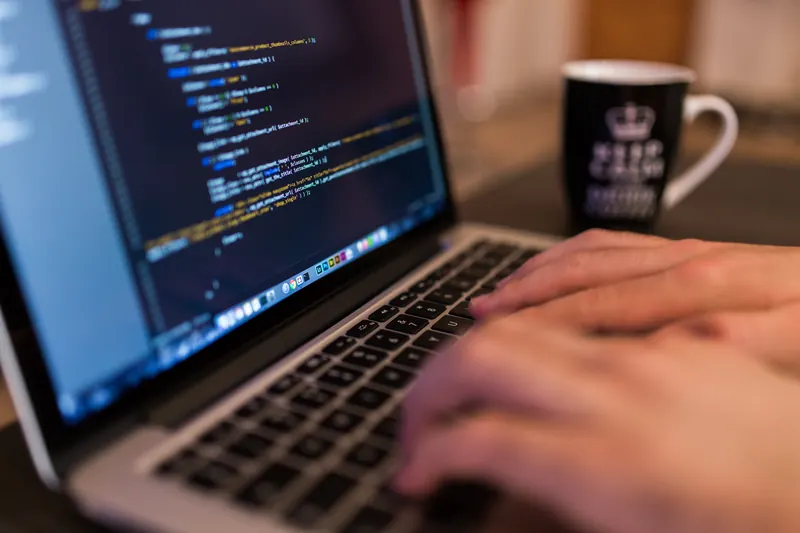 Manos escribiendo en una laptop con código en pantalla y una taza negra al fondo